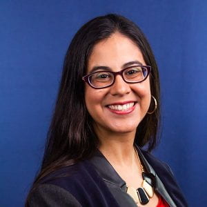 A headshot of Lorena, wearing a geometric necklace and black blazer. She has long dark hair, gold hoop earrings, and dark framed glasses. She is smiling! 