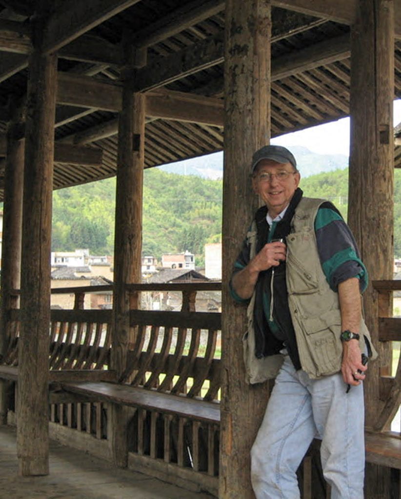 Ronald Knapp leans against a post of a covered wooden bridge with trees and buildings in the background.